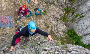 Wspinaczka podczas scala kursu. Kobieta w niebieskim kasku wspina się po skale, asekuruje ją osoba w czerwonym kasku.