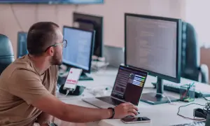 Programista aplikacji w okularach pracuje przy biurku z kilkoma monitorami i laptopem.